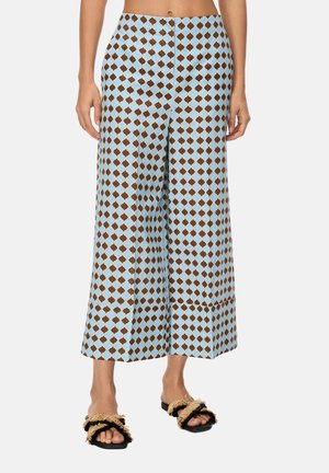 Donna che indossa pantaloni a vita alta e gamba larga con motivo geometrico blu e marrone, abbinati a sandali neri con cinturini beige con frange.