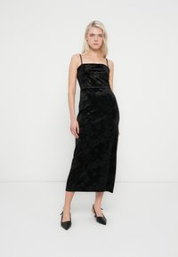 Svart fløyelskjole med blomstermønster, tynne stropper og innsvingt liv, som går over til en midilengde skjørt. Bruker svarte spisse hæler.