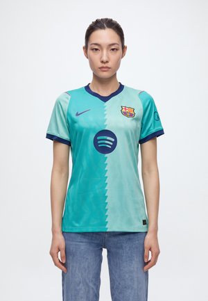 Une femme porte un maillot de football turquoise du FC Barcelone, avec des finitions bleu marine, l’écusson du club, le swoosh Nike et le logo Spotify, associé à un jean.