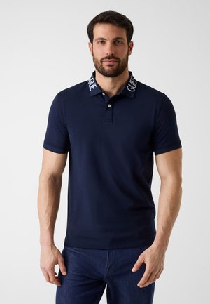 Homme aux cheveux courts et foncés avec une barbe, portant un polo bleu marine avec le texte « GUESS » sur le col et un jean bleu foncé, debout devant un fond blanc.