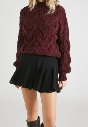 Femme portant un gros pull en maille torsadée bordeaux associé à une mini-jupe plissée noire et des bottes marron foncé.