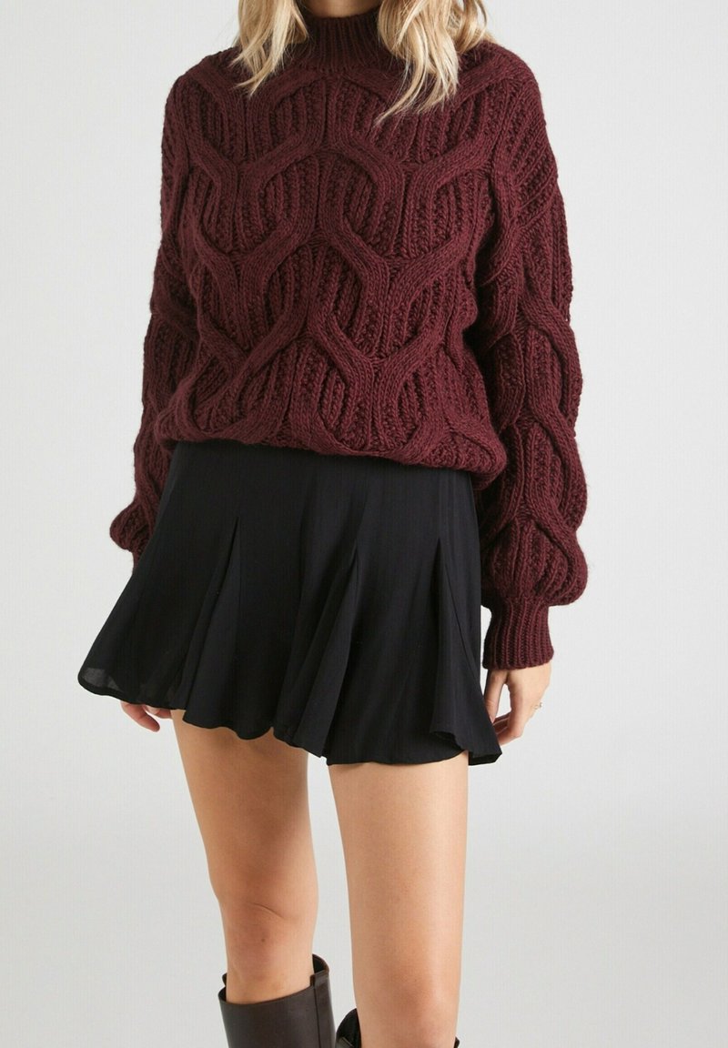 Femme portant un gros pull en maille torsadée bordeaux associé à une mini-jupe plissée noire et des bottes marron foncé.