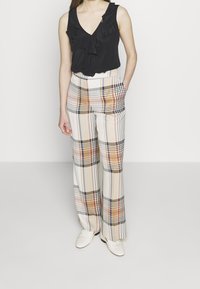 Top senza maniche nero con ruches abbinato a pantaloni a gamba larga in plaid nei colori crema, arancione e nero. Le scarpe bianche completano l'outfit.