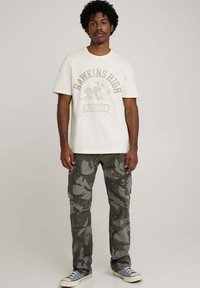 Biały t-shirt z nadrukiem "HAWKINS HIGH", zestawiony z szarymi spodniami cargo w kamuflażu oraz jasnoniebieskimi sneakersami. Casualowy, luźny krój, bez wzorów na spodniach.