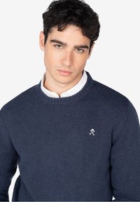 Suéter de punto azul marino con cuello redondo y puños acanalados. Presenta un pequeño logotipo gráfico blanco en el pecho. Textura suave, ajuste clásico.