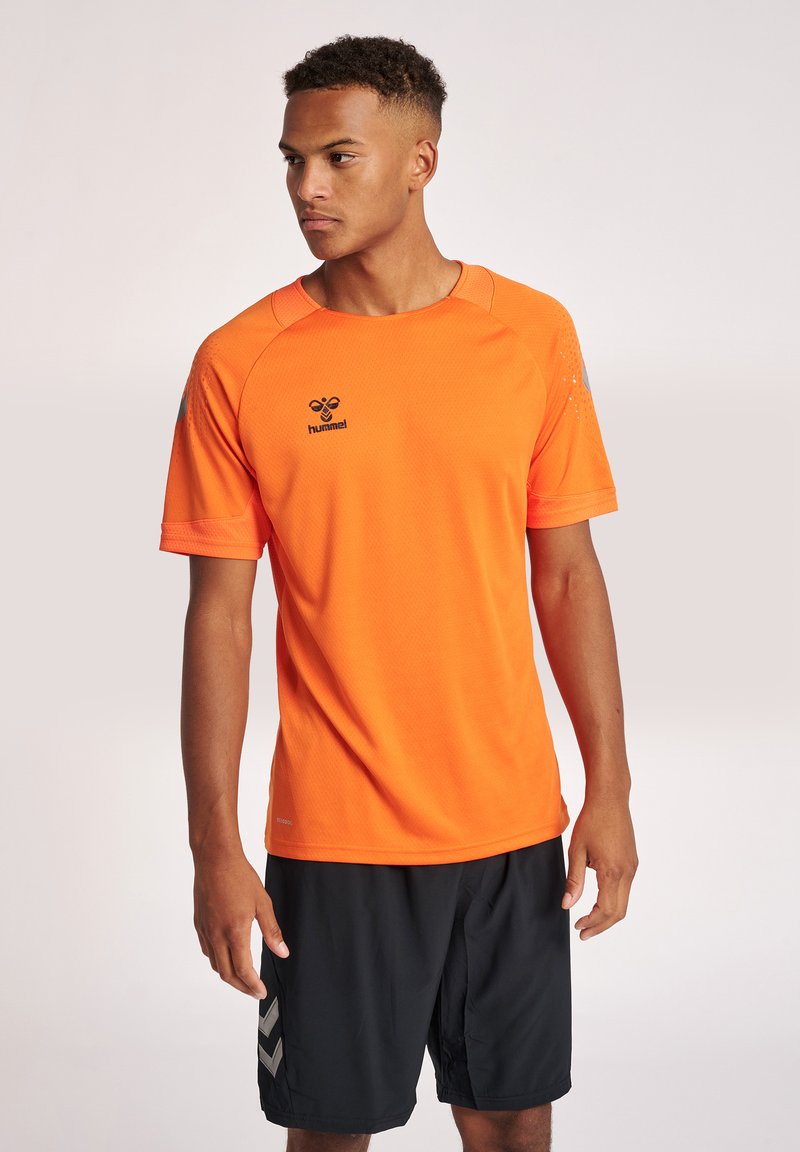 Jonge man draagt een feloranje Hummel sportshirt en zwarte sportshorts, staand tegen een effen lichte achtergrond.