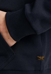 Granatowy sweter z ribbowymi mankietami i dołem. Posiada małą pomarańczową metkę z boku z napisem "FREEDOM UNLIMITED". Miękka tekstura.