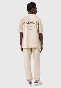 Mand iført en beige oversized T-shirt med "ALLSAINTS" og japansk tekst, matchende beige bukser og hvide sneakers med slangeskindsdetaljer.