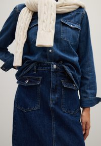Camicia di jeans con due tasche sul petto, abbinata a una gonna di jeans a vita alta con tasche frontali. Sciarpa di maglia a treccia color crema drappeggiata.