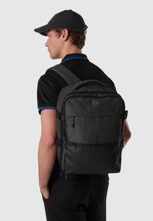 Uomo con polo nera e cappellino, che indossa uno zaino nero e grigio con molteplici scomparti con cerniera, in piedi di lato davanti a uno sfondo neutro.