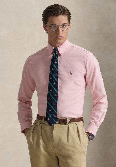 Man met ronde bril, lichtroze overhemd met knopen, patroon navy en groene stropdas, beige broek, bruine riem en polshorloge, staand met handen in de zakken.