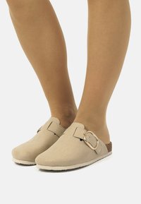 Zuecos de ante beige con una correa con hebilla, punta redondeada y suela texturizada. Diseño ligero con una plantilla de corcho para mayor comodidad.