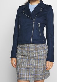 Marinblå bikerjacka i fejksmockad med dragkedjor, sidofickor med dragkedja, axelklaffar och en figursydd silhuett, kombinerad med en rutig kjol i brunt och blått.