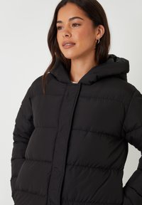 Sort puffy jakke med hætte, kviltet design, frontlukning og rummelig pasform. Glat tekstur med synlige stikninger.