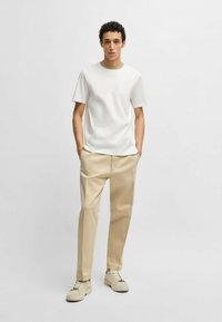 T-shirt bianca a maniche corte con colletto crema, abbinata a pantaloni beige e sneakers chiare. Il modello sta in piedi con le mani nelle tasche.