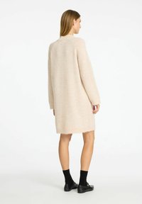 Ljusbeige stickad cardigan-klänning med mjuk textur, sänkta axlar och rak skärning, försedda med en fransad fåll.
