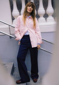 Ženska z dolgimi lasmi, naslonjena na ograjo stopnic, oblečena v svetlo roza jakno, črtasto majico, temne široke hlače in rjave čevlje.