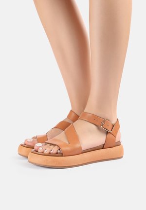 Pieds nus portant des sandales compensées en cuir marron clair avec des brides à la cheville et des boucles métalliques sur un fond clair uni.