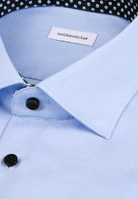 Chemise habillée bleu clair avec des boutons noirs, un col pointu, et une étiquette "seidensticker" à l'intérieur du col.