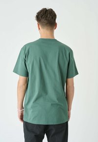 Muž s krátkými vlasy stojí čelem k obličeji, na sobě má zelené tričko s krátkým rukávem a černé kalhoty na jednoduchém světlém pozadí.