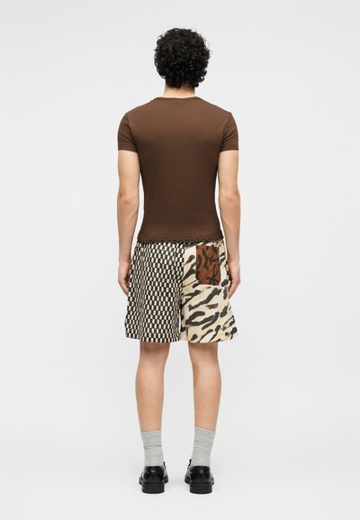 Άνδρας που στέκεται με την πλάτη προς τα πίσω, φοράει ένα καφέ εφαρμοστό κοντομάνικο πουκάμισο και σορτς με σχέδια, με μπαλώματα με καρό και ζωικά μοτίβα.