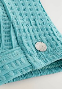 Türkiz színű, texturált anyag mintás dizájnnal. Egy ezüst kerek gomb dombornyomott "MINT VELVET" felirattal. Puha, szőtt anyag.
