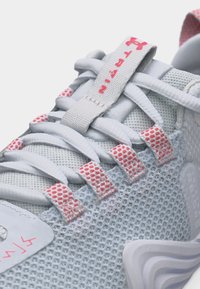 Κοντινό πλάνο λευκού αθλητικού παπουτσιού από δίχτυ με ροζ λεπτομέρειες, υφασμάτινες θηλιές για τα κορδόνια και το κείμενο «Train» στην ετικέτα της γλώσσας.