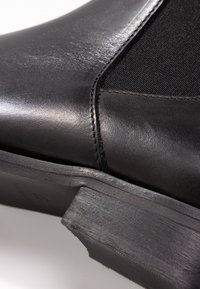 Botín de cuero negro que presenta una textura suave, panel de tela en contraste y un tacón de goma cuadrado bajo con acabado mate.