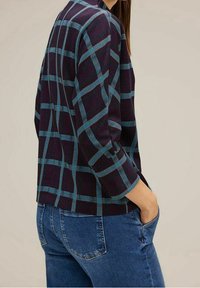 Camicetta a maniche corte a quadratini in viola scuro e teal, caratterizzata da un design boxy e un tessuto testurizzato, abbinata a jeans in denim di lavaggio medio.