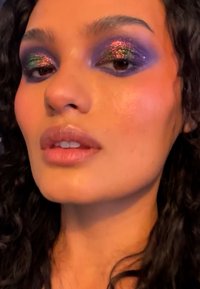 Nahaufnahme eines Gesichts mit dunklem, lockigem Haar, das auffälligen lila Lidschatten mit bunten Glitzerakzenten und natürlichen, glänzenden Lippen zeigt.