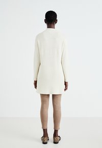 Long cardigan crème à côtes, avec des manches longues, porté sur un pantalon beige ajusté et des chaussures à imprimé léopard ; design minimaliste, sans fermetures visibles.