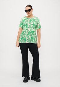 Kortärmad, grön blommönstrad topp på en ljus bakgrund, i kombination med svarta utsvängda byxor och kraftiga svarta skor. Slät textur.