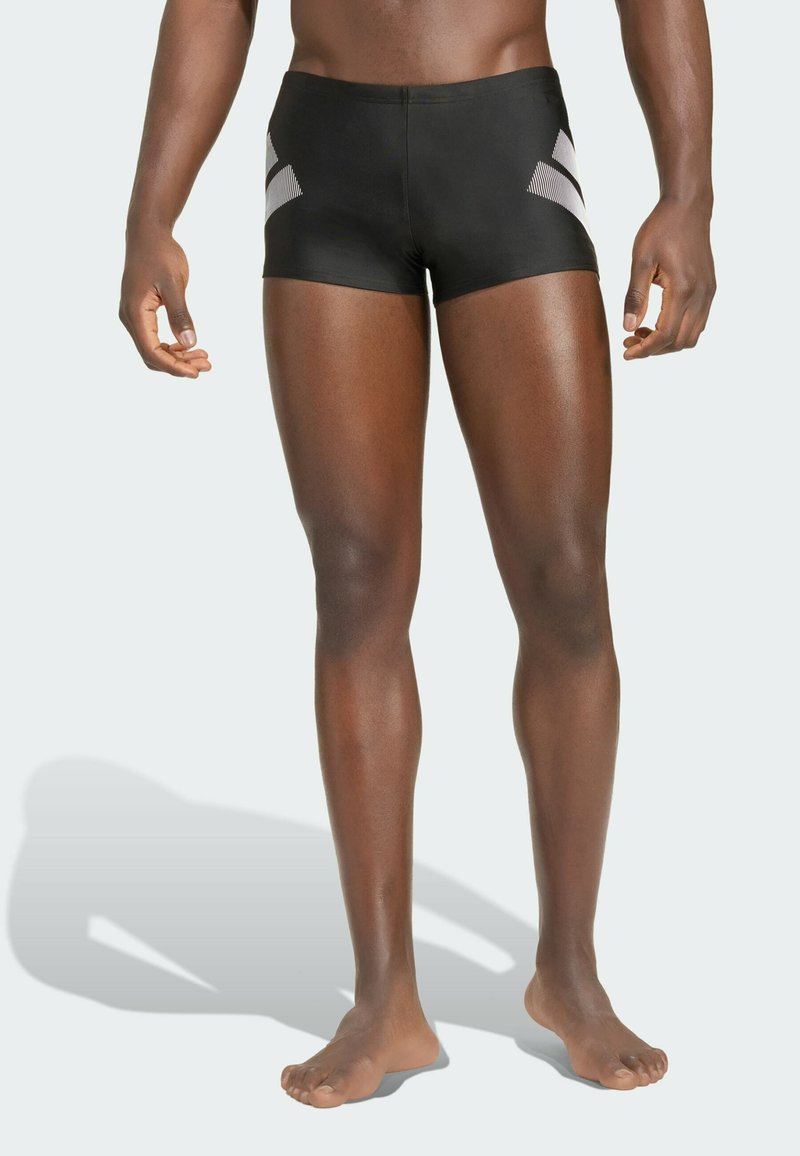 Mannequin masculin debout, pieds nus, portant un slip de bain noir moulant avec des rayures diagonales blanches sur les côtés, sur un fond neutre.