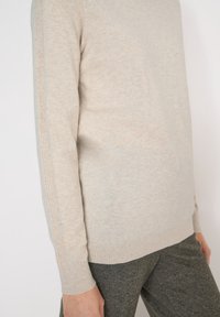 Pull en tricot beige à manches longues avec poignets côtelés et détails de couture le long des manches, porté avec un pantalon texturé gris foncé.