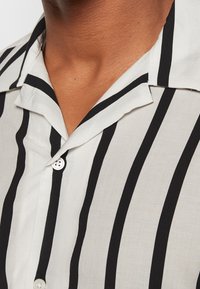 Gros plan sur le col d'une chemise à rayures verticales noires et blanches portée par une personne à la peau marron moyenne.