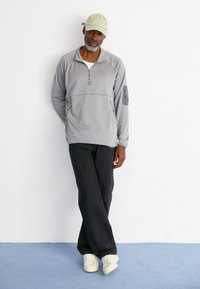 Grå hettejakke med glidelås i kragen og sidelommer; kombinert med svarte bukser og beige sneakers; grønn caps gir et komplementært preg.