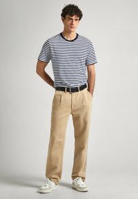 Juostuota tamsiai mėlyna ir balta marškinėliai su juodu apykaklėle, derinami su smėlio spalvos kelnėmis ir baltais sportbačiais. Modelis stovi su rankomis kišenėse.