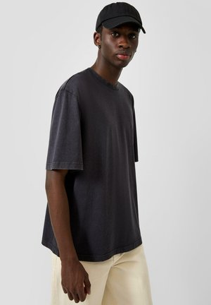 Junger Mann mit schwarzer Kappe, lockerem schwarzen T-Shirt und hellbeigen Hosen, stehend vor einem schlichten grauen Hintergrund.