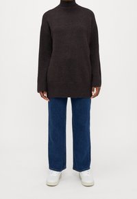 Person, der einen dunkelbraunen Rollkragenpullover, gerade blaue Jeans und weiße Sneakers trägt und vor einem schlichten Hintergrund steht.