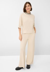 Beige, kortærmet top med sort kant ved halsen, kombineret med løstsiddende beige bukser. Modellen har sorte ankelstøvler på.