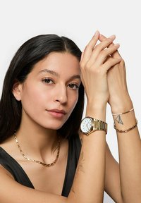 Guldklocka med en rund vit urtavla och guldkedja. Modellen bär ett guldhalsband och en annan armband, vilket visar tatueringar på hennes arm.