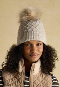 Knit wool hat in light beige with a diamond pattern, topped with a fluffy faux fur pom-pom. Worn with a beige quilted vest.