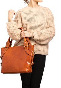 Sac fourre-tout en cuir marron avec des sangles ajustables, finition polie et matériel en métal, associé à un pull en tricot beige et un pantalon noir.