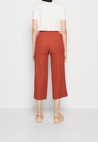 Personne portant un pantalon ample taille haute, couleur rouille, raccourci, associé à une chemise blanche à manches courtes et des sandales compensées beiges, debout.