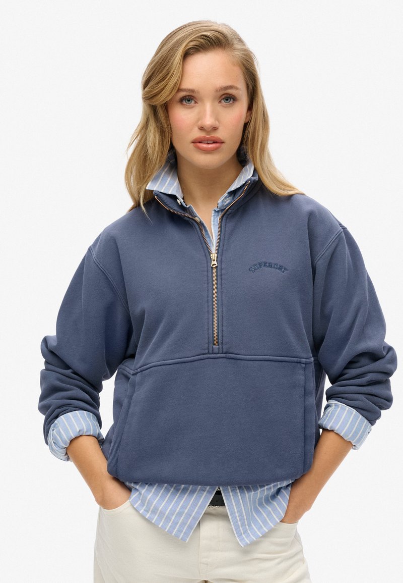 Sweat-shirt zippé en mélange de coton bleu foncé avec une fermeture éclair quart, une poche avant et un discret logo "Superdry" brodé. Superposé sur une chemise à rayures bleu clair.