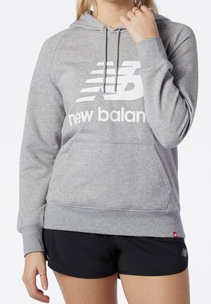 Femme portant un sweat à capuche gris New Balance avec poche avant et short de sport noir sur fond blanc.