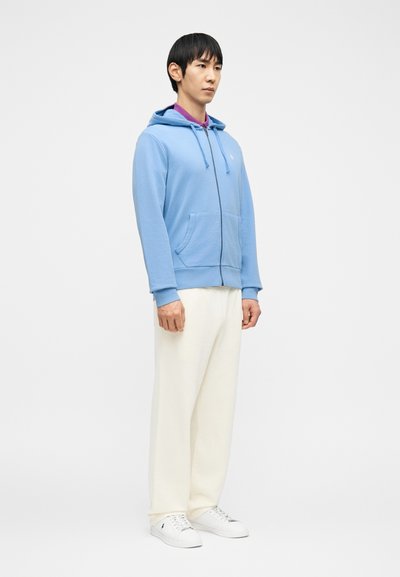 Άνδρας στέκεται φορώντας ανοιχτό γαλάζιο φούτερ με φερμουάρ, μωβ πουκάμισο, λευκό παντελόνι και λευκά αθλητικά παπούτσια μπροστά σε λευκό απλό φόντο.