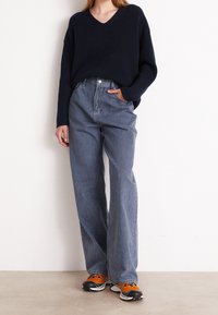 Navy ribbed knit jumper with V-neck, paired with high-waisted, loose gray jeans. The model is wearing orange and black sneakers. Minimalist background.