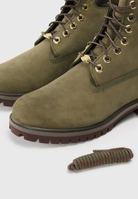 Botas de ante verde oliva con ojales dorados, suelas de goma marrón y cordones marrones. Incluye un accesorio de paracord enrollado en verde y rojo.