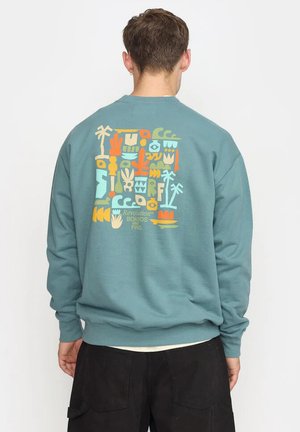 Uomo che indossa una felpa verde acqua con design tropicale astratto e colorato e la scritta "Revolution Boards & Fins" sulla schiena, abbinata a pantaloni neri.